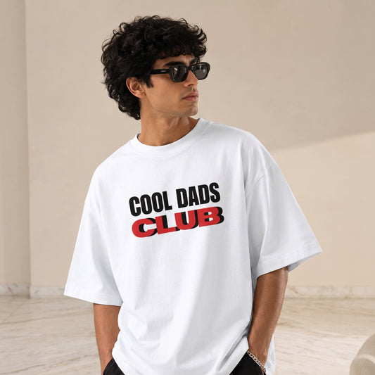 A man wearing a white oversized t-shirt with 'COOL DADS CLUB' printed on the chest in black and red, styled with black sunglasses in a minimalist beige room.